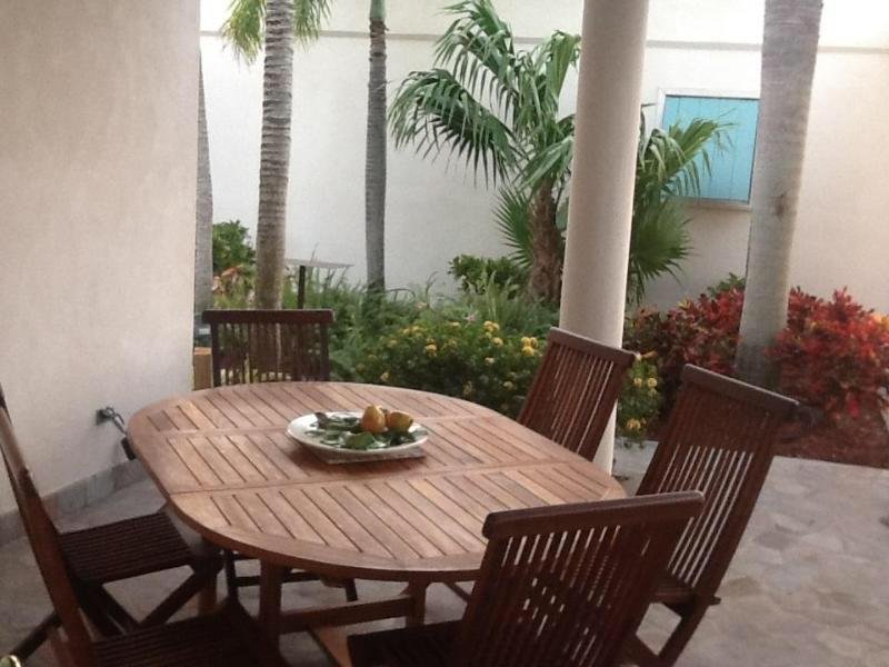 Modern outdoor patio dining area with wooden table and chairs, surrounded by lush tropical plants and palm trees, ideal for relaxing and entertaining at 17 Degrees North Realtors in Nellis AFB, Nevada.