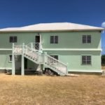 Modern two-story green house with exterior staircase and large windows, located in a sunny, tropical area ideal for real estate investments in Nevis.