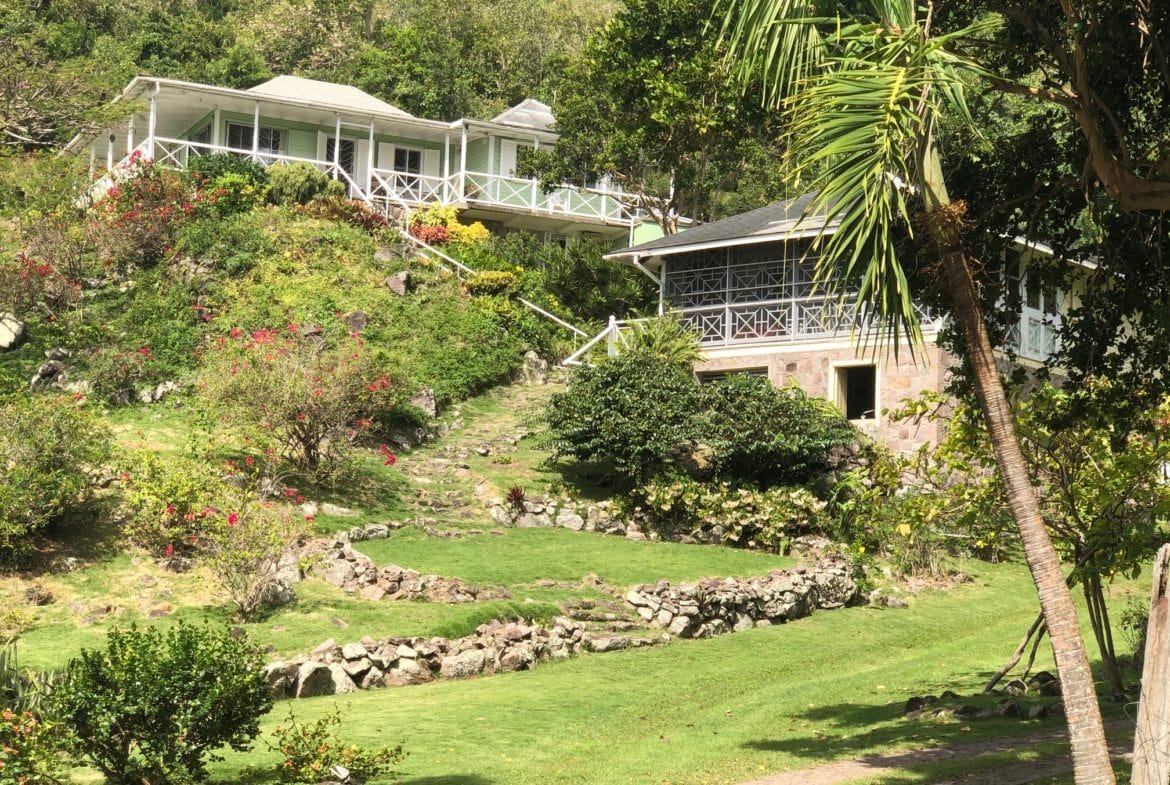 Lush tropical hillside property with scenic views, featuring charming green and white homes, surrounded by vibrant plants and trees in a serene, natural setting.