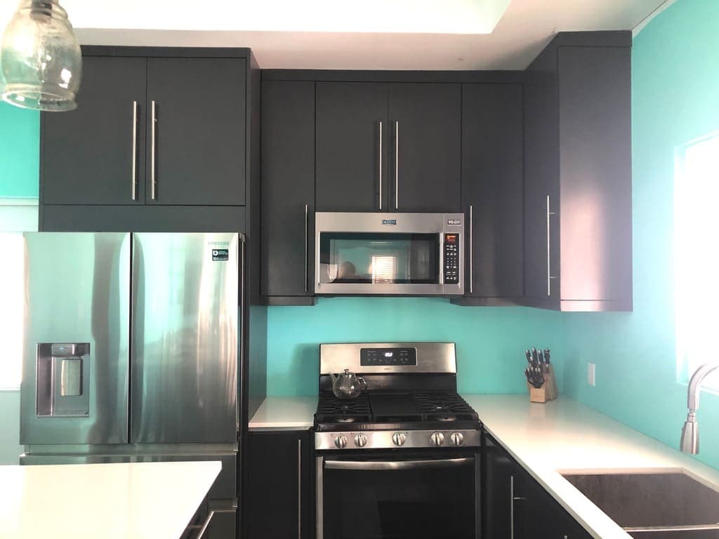 Stainless steel refrigerator, microwave, and gas stove in modern kitchen with turquoise wall and black cabinetry.