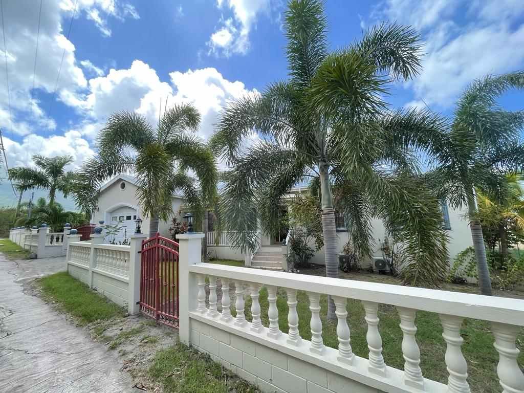 Bright white tropical style house with palm trees on a sunny day, surrounded by a decorative white fence and red gate, located in a peaceful area with blue skies and fluffy clouds.