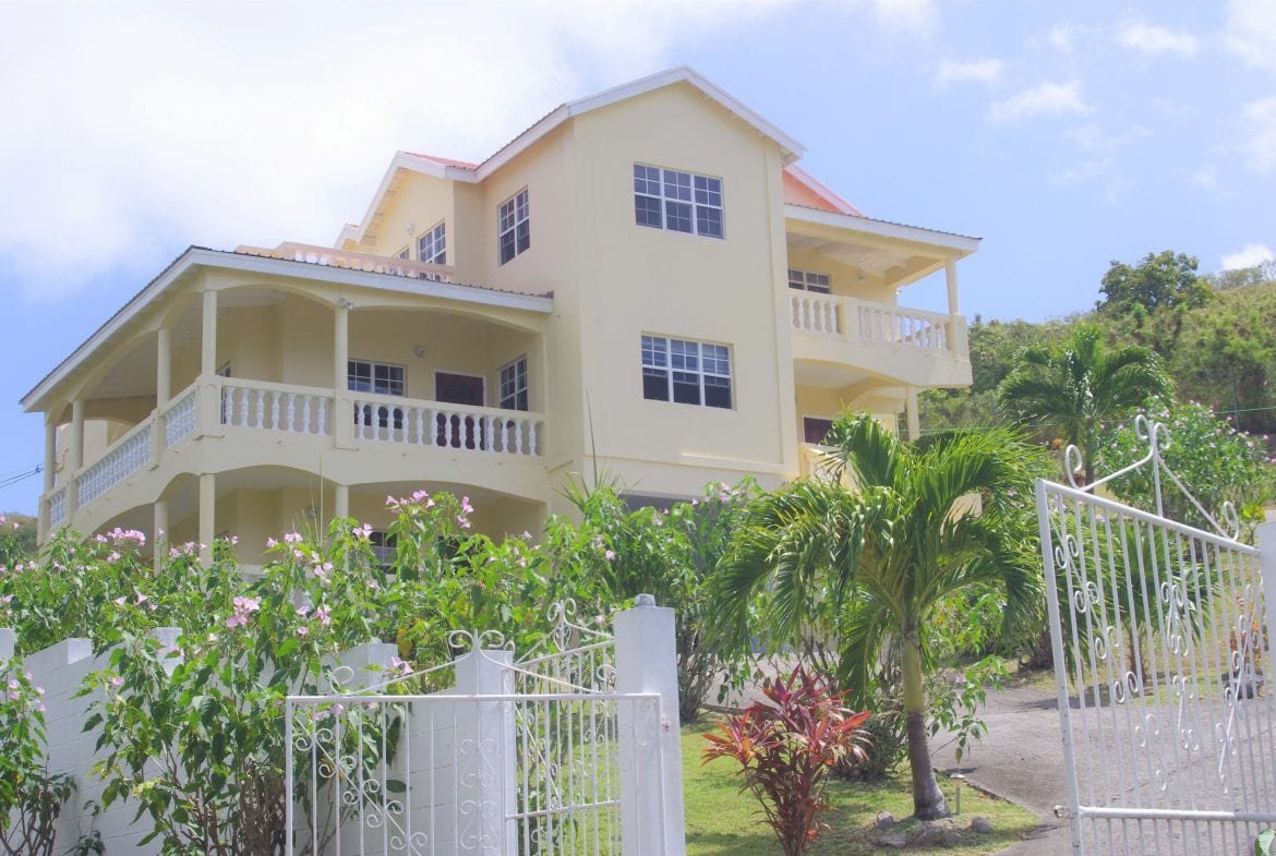 Stylish multi-story yellow house with wide balconies, surrounded by lush tropical landscaping, located in a scenic hillside area in Nevis, ideal for luxury real estate investment.