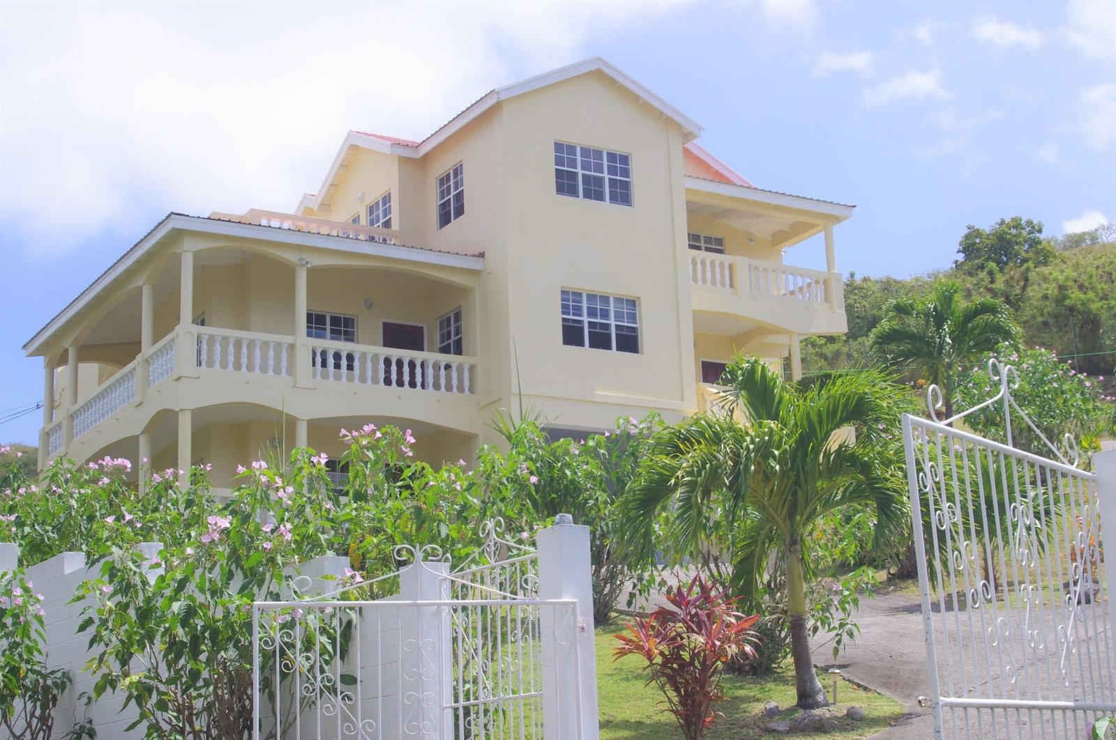 House for sale pic 3 Stylish multi-story yellow house with wide balconies, surrounded by lush tropical landscaping, located in a scenic hillside area in Nevis, ideal for luxury real estate investment.