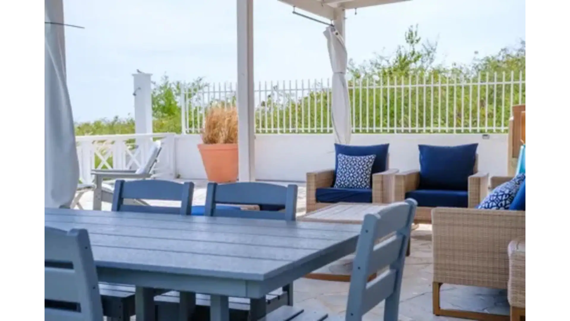 Modern outdoor patio with comfortable seating and dining area in a beachside property, perfect for relaxing and entertaining. Featuring wicker chairs, blue cushions, and a scenic view of greenery.