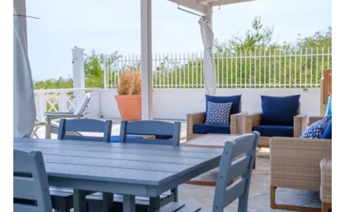 Modern outdoor patio with comfortable seating and dining area in a beachside property, perfect for relaxing and entertaining. Featuring wicker chairs, blue cushions, and a scenic view of greenery.